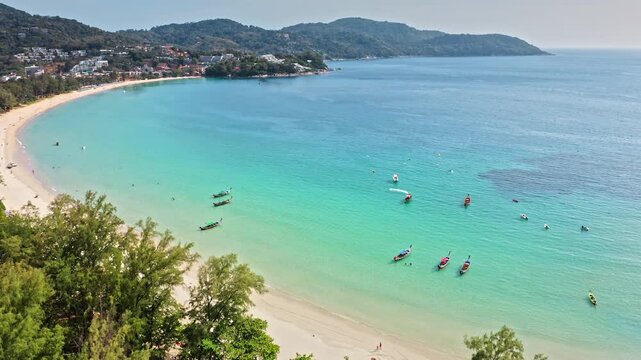 Kata Beach, Phuket, expansive tropical bay with turquoise water, white sand, longtail boats near shore and lush green hills under clear blue sky, tranquil island. Summer travel paradise. Aerial flight