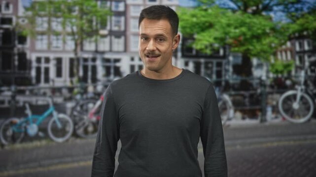 Man points finger while smirking with visible moustache on street by parked bicycles and canal houses; amusement irony.