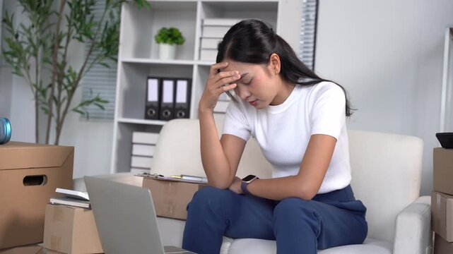 Overwhelmed Entrepreneur: A young businesswoman sits at her desk, head in hand, looking stressed and overwhelmed by her work, surrounded by stacks of boxes and a laptop.