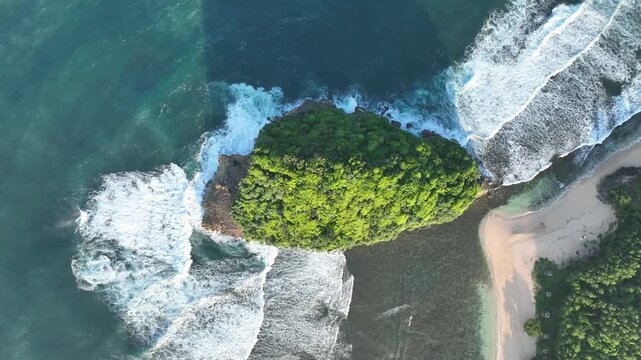 Aerial view of Pantai Ngudel featuring a lush green rocky outcrop surrounded by turquoise ocean waves and a sandy beach in Gedangan, Jawa Timur, Indonesia.