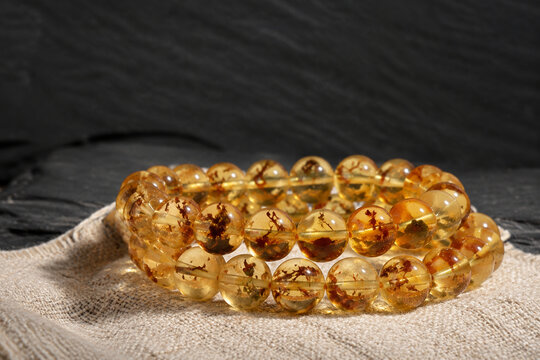 Honey-colored resin bead bracelet on linen cloth, organic plant inclusions, dark slate background, amber tones, jewelry presentation