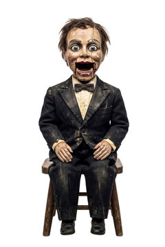 Creepy cracked ventriloquist dummy in a dusty black suit sitting on a stool isolated on transparent background.