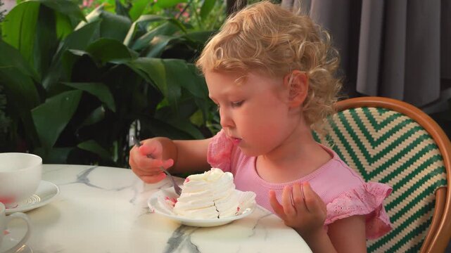 Medium close up of a lovely Caucasian girl, about four and a half years old with blonde curly hair, sitting at an outdoor cafe table, focused on eating a decadent Pavlova ice cream dessert with fresh 