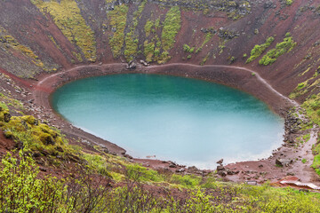 The crater lake lies within layered red scoria and green moss at Kerid Crater, Iceland. An elevated rim perspective reveals blue water, wet volcanic slopes, and curved shoreline after rain. © Artem