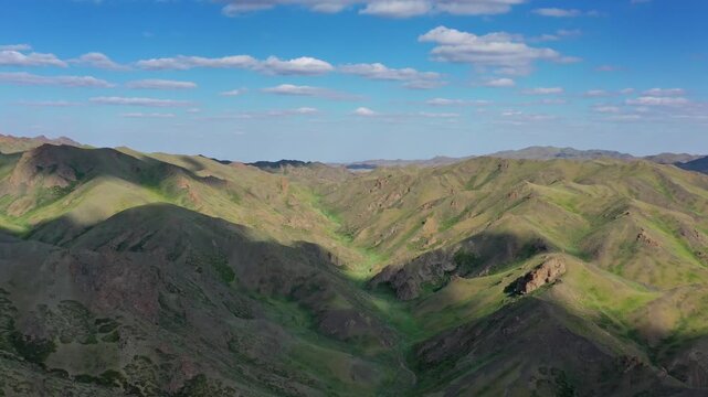 Aerial view of rolling green hills and mountains under a blue sky with scattered clouds in Mongolia.