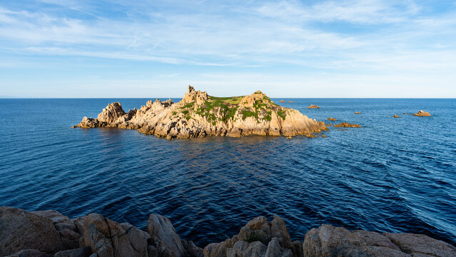 The rocky islet Rocher des Portes surrounding deep blue sea waters off the coast of Cap Camarat near Ramatuelle, France. Bright sunny daylight accentuates the coastal rock and vibrant landscape.