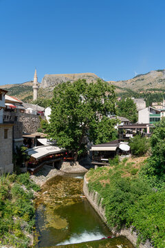 Mostar, Bosnia and Herzegovina - 22 June 2025: View of the Radobolja stream in the historic old town, with stone buildings, a minaret, lush trees, and restaurant terraces.