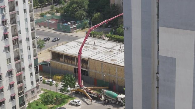 High-angle view of workers pouring a rooftop slab with a concrete boom pump between apartment buildings while traffic passes close to the active urban worksite.