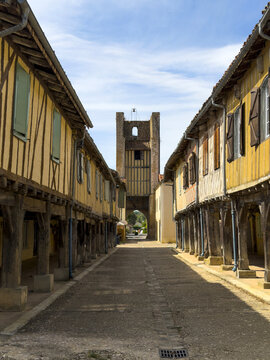 Arcaded village street with hanging flower baskets and a distant bell tower, Gers, Tilliac, France