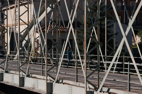 Person walks across metal truss bridge. Sunlight filters through steel beams. Background shows buildings and greenery. Structure has classic industrial design