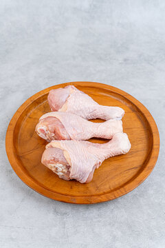 Three raw fresh chicken drumsticks on a round wooden plate against a light grey concrete background with copy space