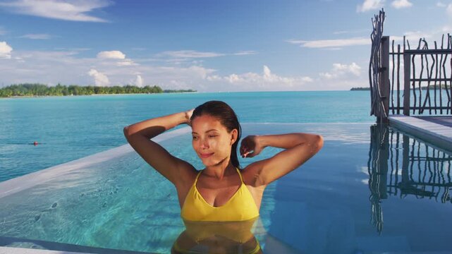 Luxury swimming pool Asian bikini model woman wearing yellow fashion swimsuit relaxing in infinity pool at overwater bungalow resort, tropical vacation. Raw video of real woman model without makeup