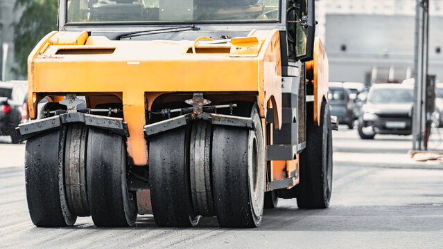 A heavy-duty roller works on a city street, compacting the asphalt as cars pass by. The machine effectively prepares the surface