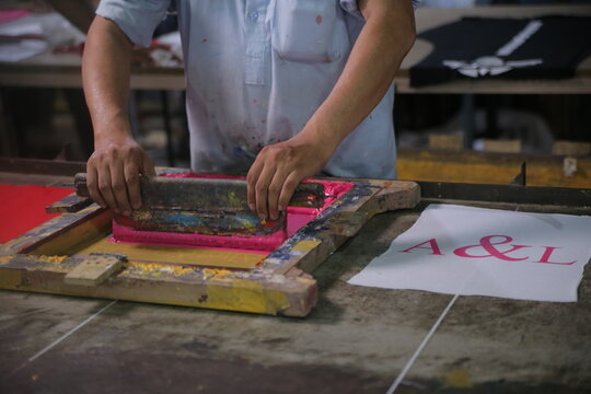Lahore, Pakistan - 02 October 2024: View of a worker using a squeegee to apply pink ink through a screen onto fabric in a textile workshop at Quaid-e-Azam Industrial Estate.