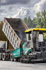 A dump truck is unloading material into a paving machine at a construction site. The heavy equipment is actively engaged in road construction © Anoo