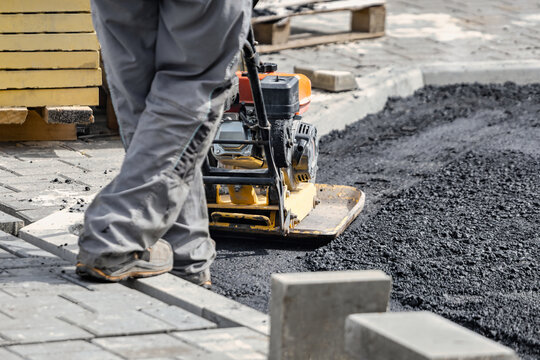 A person operates a compactor machine to flatten freshly laid asphalt on a busy street. Nearby, stacks of materials are organized for the work