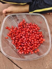 Top-down view of fresh, juicy red pomegranate seeds in a clear plastic bowl on a textured stone floor, with a child's bare foot visible in the background representing healthy everyday snacking. © Lovely