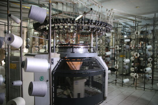 Lahore, Pakistan - 02 October 2024: View of a circular knitting machine in operation at the Quaid-e-Azam Industrial Estate, surrounded by numerous white yarn spools in a textile factory.