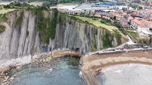 Cinematic aerial drone footage flying backward and panning left, revealing the coastal town of Zumaia nestled behind the dramatic vertical flysch rock formations of Itzurun Beach, Spain &ndash; 2026