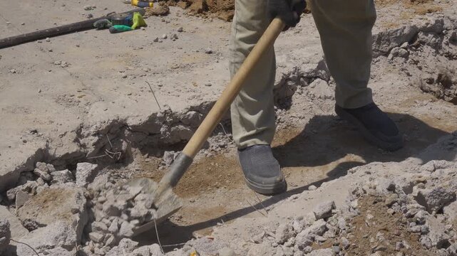 A cropped construction worker shovels heavy concrete rubble from demolished pavement while exposed wire and unstable debris create workplace hazards during outdoor road repair.
