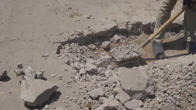 Close-up of an unrecognizable worker shoveling broken concrete and gravel during road demolition as exposed wire and casual footwear highlight unsafe working conditions.