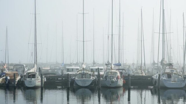Sailboats and motor yachts moor at harbor dock in dense coastal fog. Tall masts reflect in calm water on hazy morning. Quiet marina provides safe boat storage and maritime travel background.