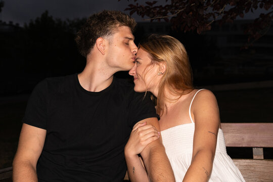 A couple is sitting on a bench, and he kisses her on the forehead