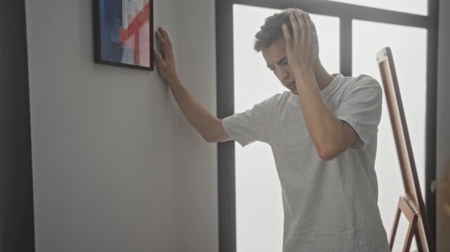 Young man with hand on forehead, leaning on wall and supporting himself by an easel in studio; distress dizziness sickness.