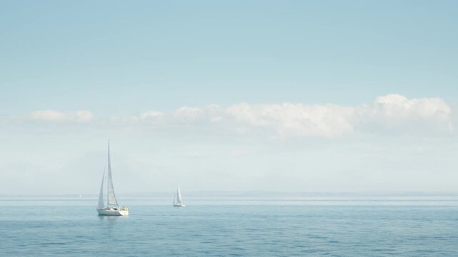 Two sailboats sail across the calm blue sea water under a cloudy sky. White yachts glide smoothly along the ocean on a summer voyage. Nautical travel inspires freedom and peaceful leisure.
