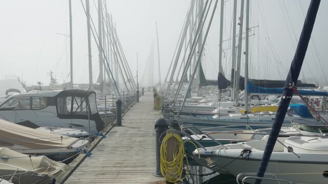 Sailboats dock along a wooden marina pier on a foggy morning. Moored yachts line the walkway in dense mist at the harbor. This boardwalk provides quiet yacht berthing and secure boat mooring.