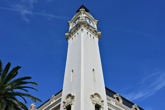 white stucco clock tower. the Port Authority of Valencia, Spain. beach area. large round white clocks. black handles. lush green palm tree. narrow vertical window. blue sky. travel , tourism concept 