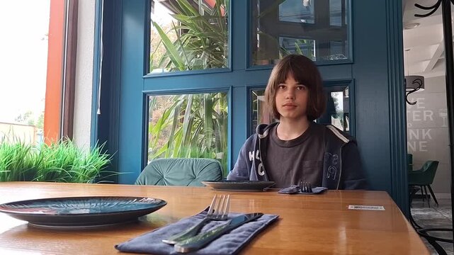13 year old boy sits at a table in a restaurant waiting for food in the afternoon