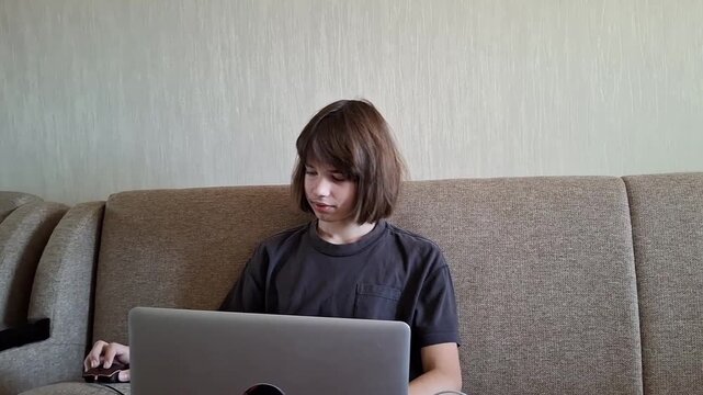 13 year old boy sits on sofa and focuses on laptop screen while using computer in living room