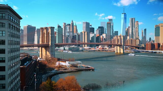 New York, USA, 1 April 2026 : Brooklyn Bridge spanning East River toward Lower Manhattan. Ferries cross beneath the suspension towers as skyscrapers rise behind a waterfront carousel pavilion.