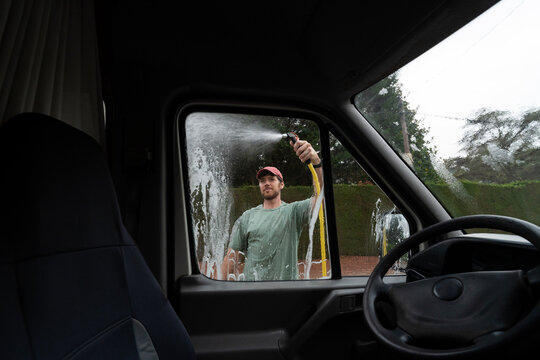 Man Washes Van Window With Hose in Driveway