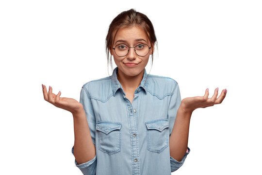 Hesitant uncertain woman shrugs shoulders, has displeased unaware expression, cant make choice between two items, wears round spectacles, models against pink background. Who cares in this situation?