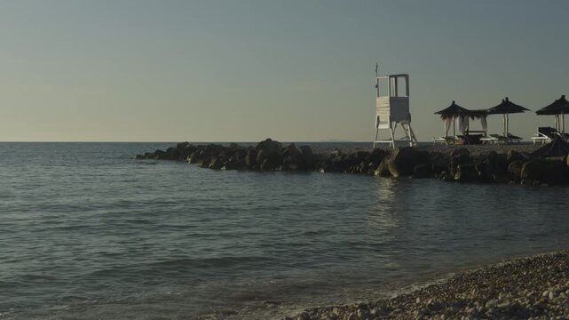 Wooden Lifeguard Tower Standing On Stone Breakwater Pier Beside Sun Umbrellas On Quiet Coastline Beach. Resort Vacation