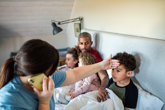 Mother checks sick son's fever in bed at home while calling doctor