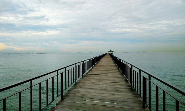 Long wooden jetty pier stretching into calm tropical ocean under a soft cloudy sky at peaceful evening