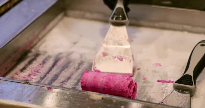 Pink berry ice cream roll being prepared with metal spatula on frozen plate, traditional Thai street food dessert making process, commercial kitchen background.