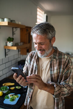 Smiling middle-aged man using smartphone while preparing healthy food at home kitchen