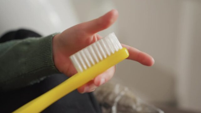 Close up of person holding and rotating yellow toothbrush with white bristles showing both sides. hand is wearing green long sleeve shirt against indoor background with soft lighting