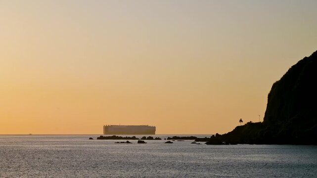 A large cargo ship sails on the calm ocean horizon during a golden sunrise, viewed from the rocky coast of Dawulun Beach in Keelung, Taiwan.