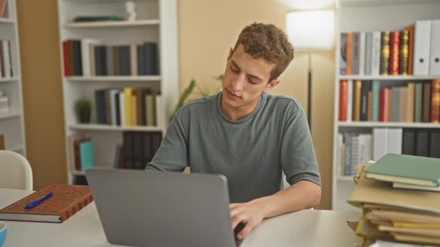 Young man with laptop and books, hands behind head stretching at a studyroom with bookshelves, lamp and stacked notebooks; study break relaxed.