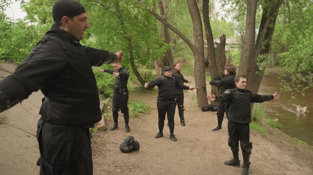 Close up of group practicing martial arts outdoors under instruction. Participants wear protective gear and maintain synchronized stances near trees with water in background. Soft natural