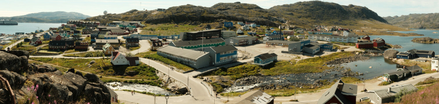 Qaqortoq, Greenland - 27 August 2025: View of colorful houses in Qaqortoq nestled among rocky hills, with a stream flowing through the town and coastal landscape under a clear sky.