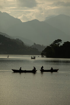 Pokhara, Nepal - 31 July 2026: View of people rowing wooden boats on Phewa Lake with the Himalayan mountains in the background under a cloudy sky.