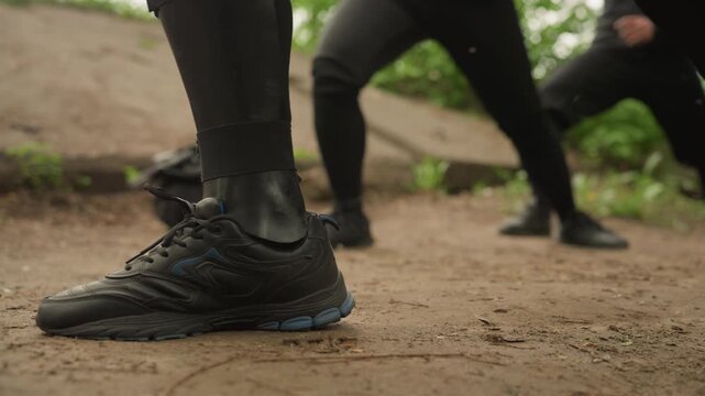Close up of feet in black athletic shoes performing leg stretches on dirt ground with green foliage background. Footwear details like laces and sole texture are visible showing movement