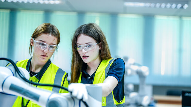 Two female robotics engineers in an R and D lab test an artificial intelligence cobot. They are smiling, inspecting the advanced prototype hardware, sensors, and programming.