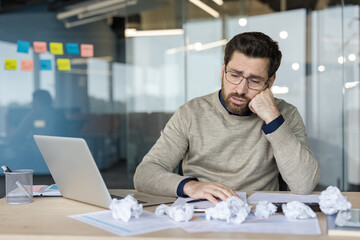 Obraz premium Tired businessman slumped at office desk, overwhelmed by workload and burnout, surrounded by crumpled papers and documents while struggling with laptop and deadlines